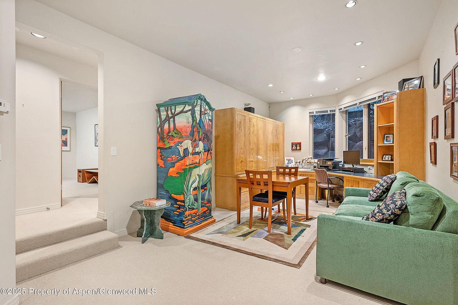 This is an interior shot of a home office or study area. The room features a unique painted cabinet, a wooden desk and chair set, and a comfortable green sofa. There's also built-in cabinetry and shelving, along with a window providing natural light, creating a functional and inviting workspace.