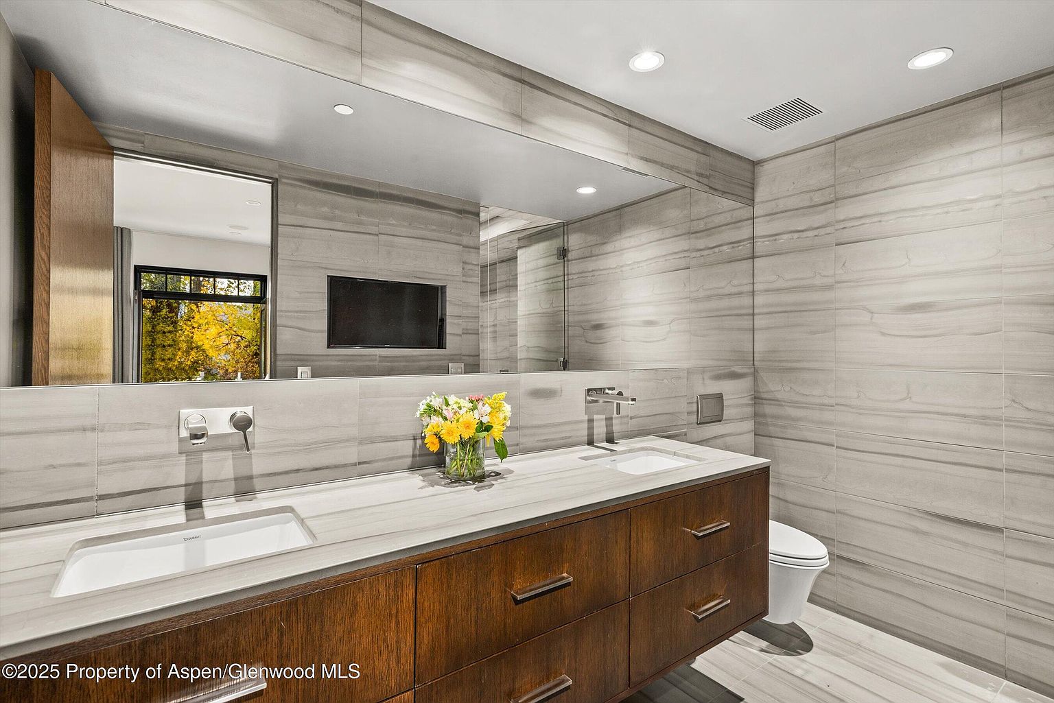 This is a modern primary bathroom featuring a double vanity with a light-colored countertop and dark wood cabinetry. The walls are covered in horizontal gray-toned tiles, and a large mirror spans the length of the vanity. A wall-mounted toilet is visible to the right, and a vase of flowers adds a touch of color to the space.