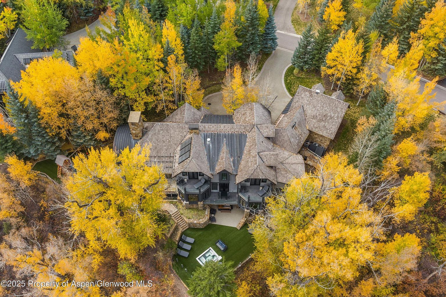 This aerial view showcases a luxurious home nestled among vibrant autumn foliage. The house features a complex roofline with multiple gables and dormers, possibly with solar panels, and a stone chimney. A well-manicured lawn with outdoor seating and a fire pit adds to the property's appeal, while a winding driveway leads to the residence, surrounded by a mix of evergreen and deciduous trees in full fall color.