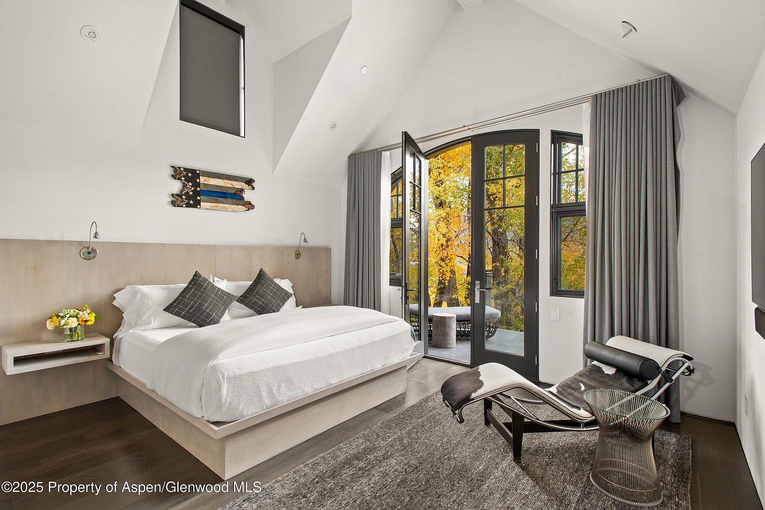 This is a primary bedroom featuring a large bed with a light wood frame and headboard, adorned with white linens and gray patterned pillows. The room has an open feel with access to a balcony through black-framed glass doors, framed by gray curtains. A modern chaise lounge and wire side table add to the room's contemporary style, complemented by a gray rug and dark wood flooring.