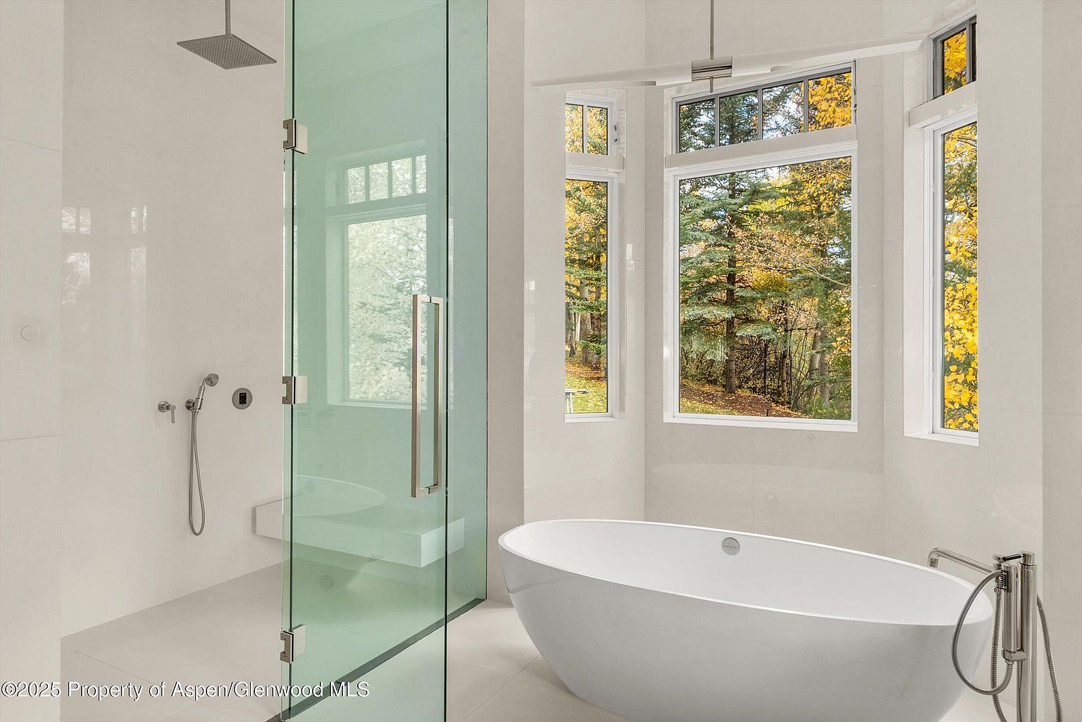 This is a bright and modern primary bathroom featuring a glass-enclosed shower with a built-in bench and a rainfall showerhead. A freestanding oval bathtub sits near a large window offering a view of trees with autumn foliage. The bathroom is finished with white tile and sleek fixtures, creating a spa-like atmosphere.