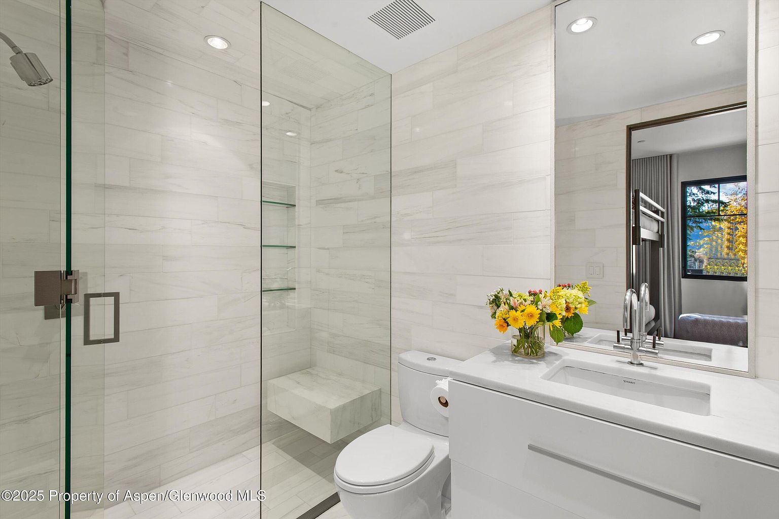 This is a bright and modern bathroom featuring white marble-like tiling throughout the shower and walls. The shower has a glass enclosure with a built-in bench and glass shelving. A white vanity with a modern sink and a toilet are visible, complemented by a large mirror reflecting a glimpse of another room, creating a clean and luxurious atmosphere.