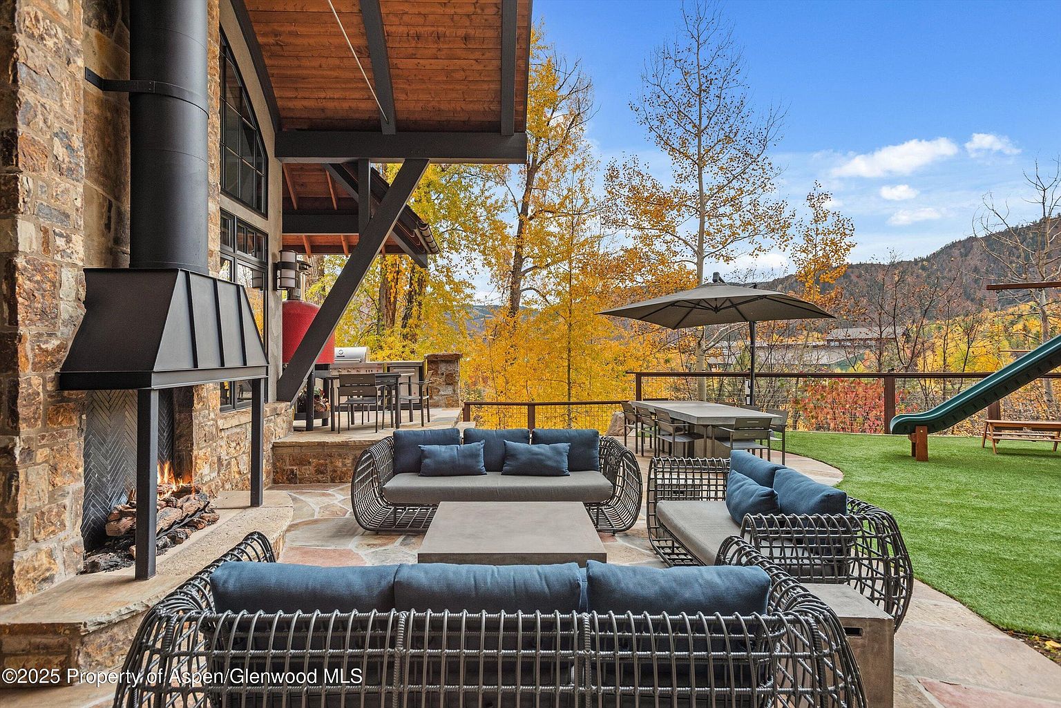 This image showcases a luxurious outdoor patio area, featuring comfortable wicker seating with plush blue cushions arranged around a stone coffee table. A stone fireplace adds warmth and ambiance, while a dining area with an umbrella and a slide in the background suggest family-friendly entertainment. The patio overlooks a scenic landscape with colorful autumn foliage, creating a serene and inviting atmosphere.