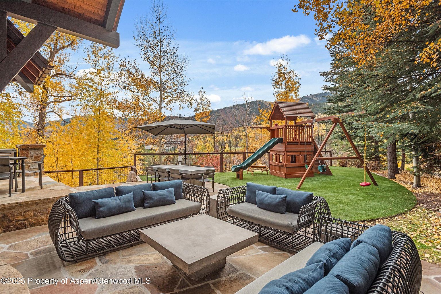 This image showcases a luxurious outdoor patio area, complete with comfortable wicker seating arrangements and a modern concrete coffee table. The patio overlooks a beautifully landscaped yard featuring a wooden playset, with a backdrop of vibrant autumn foliage and distant mountains. The scene exudes a sense of relaxation and upscale outdoor living.