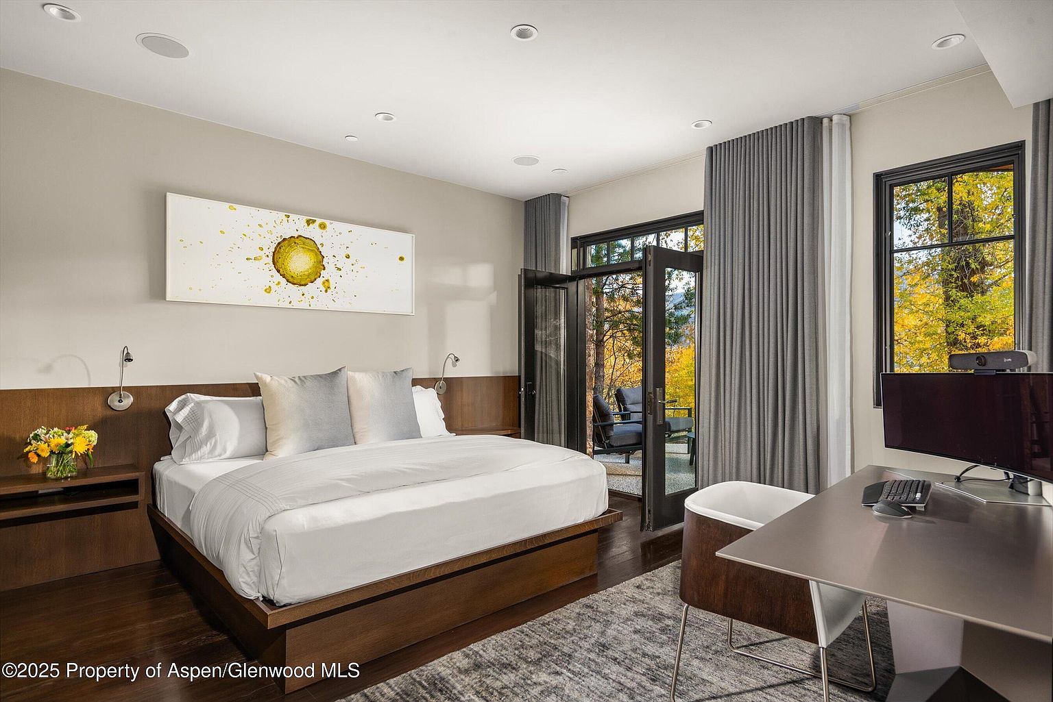 This primary bedroom features a modern design with a low-profile platform bed, a striking abstract art piece above the headboard, and dark wood flooring. The room opens to an outdoor patio through glass doors framed by elegant gray curtains, and a dedicated workspace with a sleek desk and chair is positioned near a window offering natural light. The overall impression is one of sophisticated comfort and functionality.