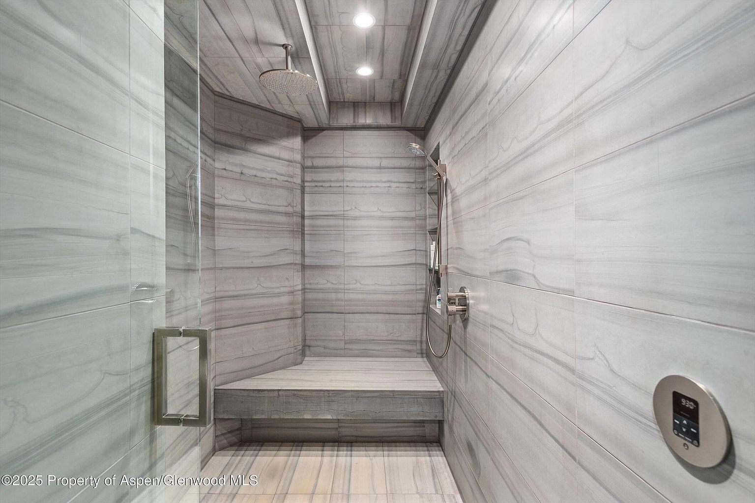 This is a modern primary bathroom featuring a walk-in shower with gray and white horizontally-laid marble-like tiles. The shower includes a built-in bench, a rainfall showerhead, and a handheld shower fixture. A glass door encloses the shower area, and a digital control panel is visible on the wall, suggesting a high-tech, luxurious experience.