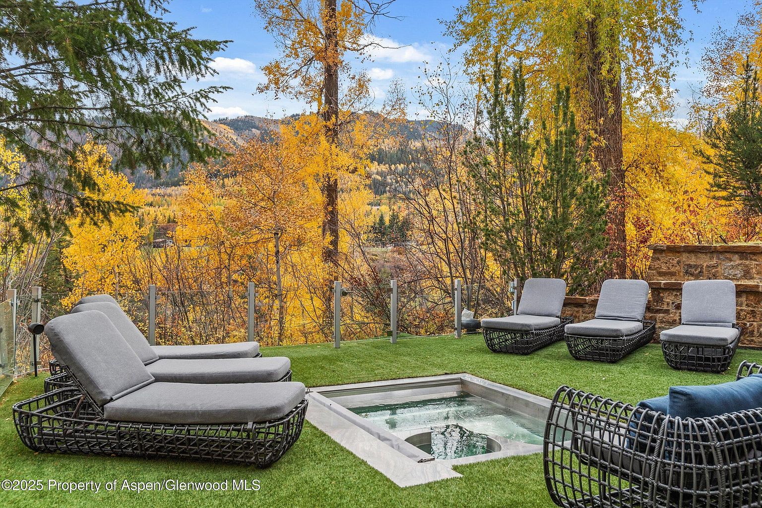 This image showcases a luxurious outdoor patio area with a built-in hot tub and comfortable lounge seating. The patio is surrounded by lush greenery and trees with autumn foliage, creating a serene and private atmosphere. The space is designed for relaxation and entertainment, offering a seamless blend of comfort and natural beauty.