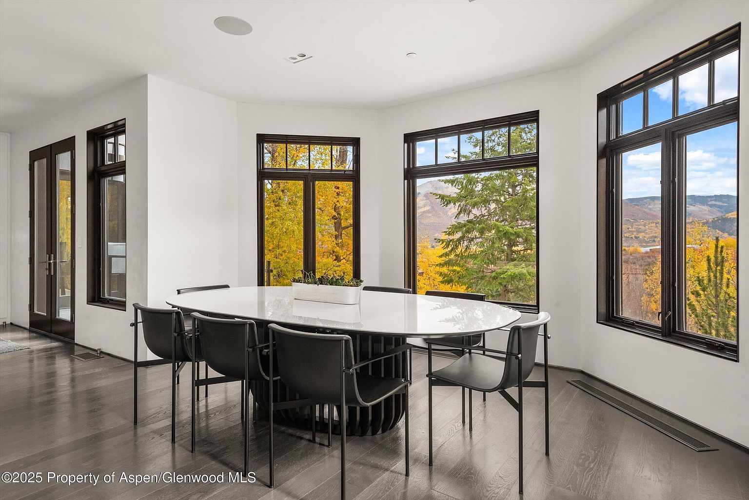 This is an interior shot of a dining room featuring a modern oval table with a white top and a unique, dark-toned base, surrounded by black chairs. Large windows with dark frames offer views of the autumn foliage outside, creating a bright and inviting atmosphere. The dark hardwood floors add a touch of elegance to the space.