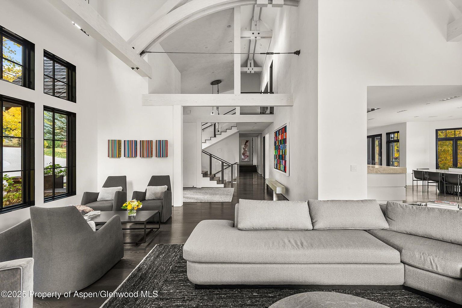 This is an interior shot of a modern living room featuring high ceilings, large windows, and an open floor plan. The room is furnished with gray sofas and armchairs, complemented by contemporary artwork on the walls. The space flows seamlessly into a hallway with stairs, creating a sense of spaciousness and luxury.