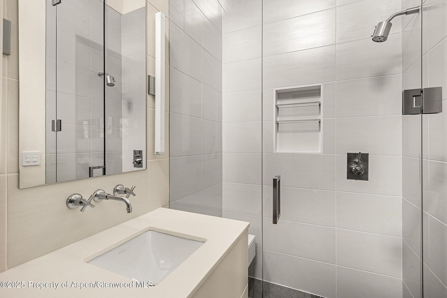 This is a well-lit bathroom featuring a modern design with light-colored, horizontally laid tiles. The bathroom includes a vanity with a white countertop and a rectangular sink, complemented by a wall-mounted faucet and a large mirror with integrated lighting. A glass-enclosed shower with a built-in niche and sleek hardware adds to the contemporary aesthetic.