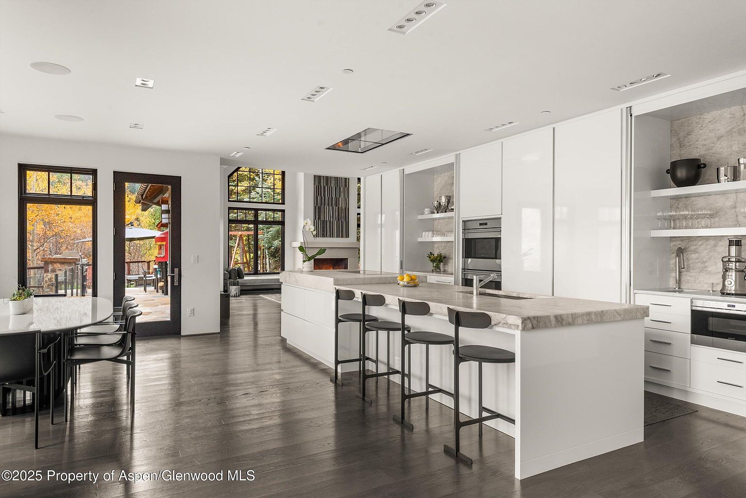 This is a bright, modern kitchen featuring sleek white cabinetry and a large island with a light-colored countertop and bar stools. The kitchen is open to a dining area and has a view of the outdoors through large windows and a door. The dark hardwood floors add a touch of warmth to the contemporary design.