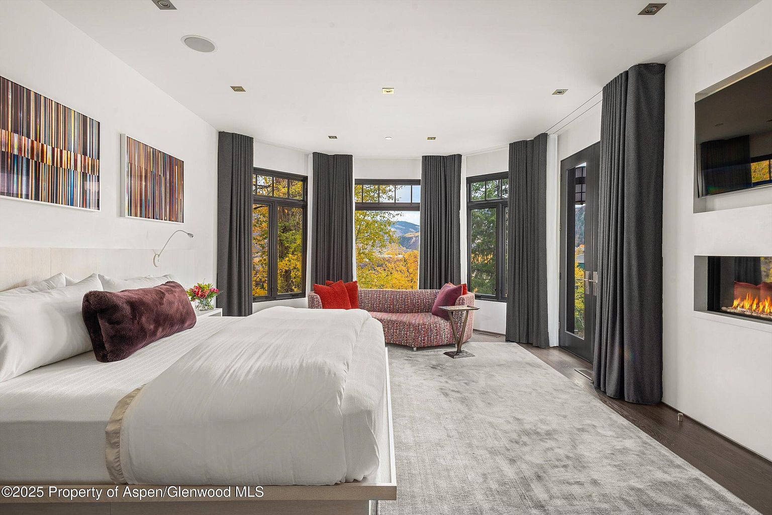 This is a luxurious primary bedroom featuring a king-size bed with white linens and a plush brown pillow. The room has a sitting area with a colorful sofa and a view of the outdoors through large windows with dark gray curtains. A modern fireplace and artwork add to the room's sophisticated ambiance.