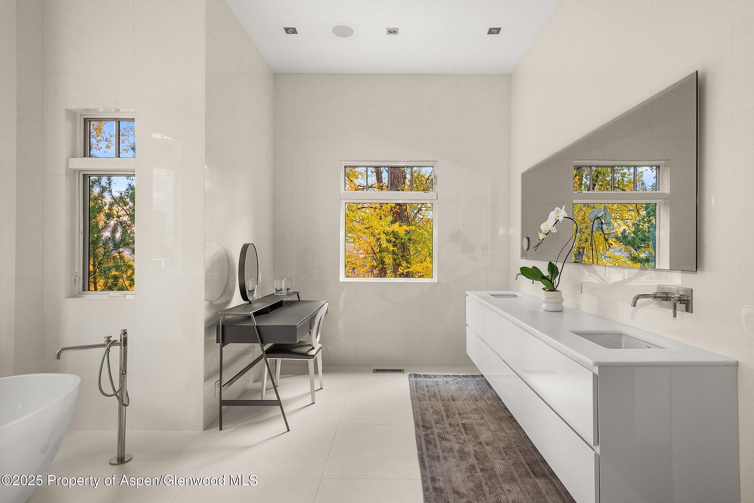 This is a bright and modern primary bathroom featuring sleek white tile walls and flooring. The room includes a floating double vanity with a large, uniquely shaped mirror, a makeup vanity with a round mirror, and a freestanding bathtub with a floor-mounted faucet. Natural light floods the space through two windows, offering views of the outdoors.