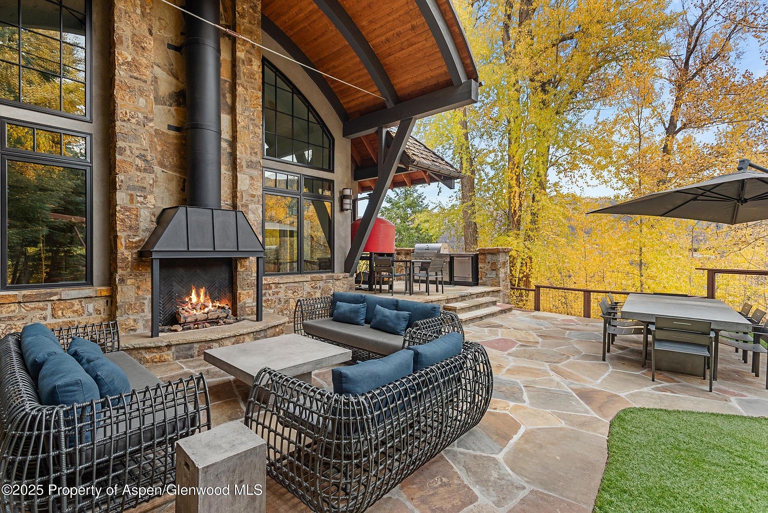 This is a beautifully designed outdoor patio area, featuring stone flooring, comfortable wicker seating with blue cushions, and a stone fireplace. The space includes a dining area with a large table and umbrella, as well as a built-in grill area, perfect for entertaining. The surrounding landscape showcases mature trees with golden foliage, adding to the luxurious and inviting atmosphere.