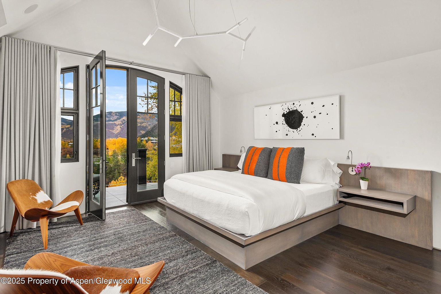 This primary bedroom features a modern design with a platform bed, minimalist artwork, and a view of the mountains through large glass doors. The room is decorated in neutral tones with pops of orange, creating a serene and stylish atmosphere. The hardwood floors and area rug add warmth and texture to the space.