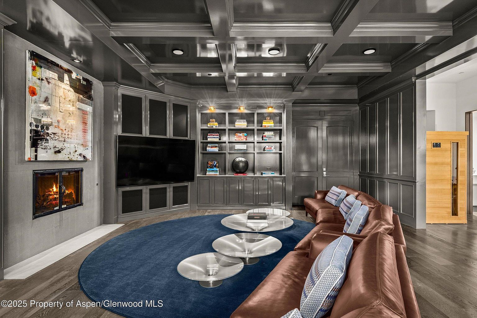 This is an interior shot of a luxurious living room featuring a dark gray color scheme with a coffered ceiling and recessed lighting. A large, curved brown leather sofa with patterned pillows sits on a round blue rug, complemented by three round glass coffee tables. A fireplace with a modern art piece above it adds a focal point, while built-in shelving and a wooden sauna are visible in the background, creating a sophisticated and comfortable atmosphere.