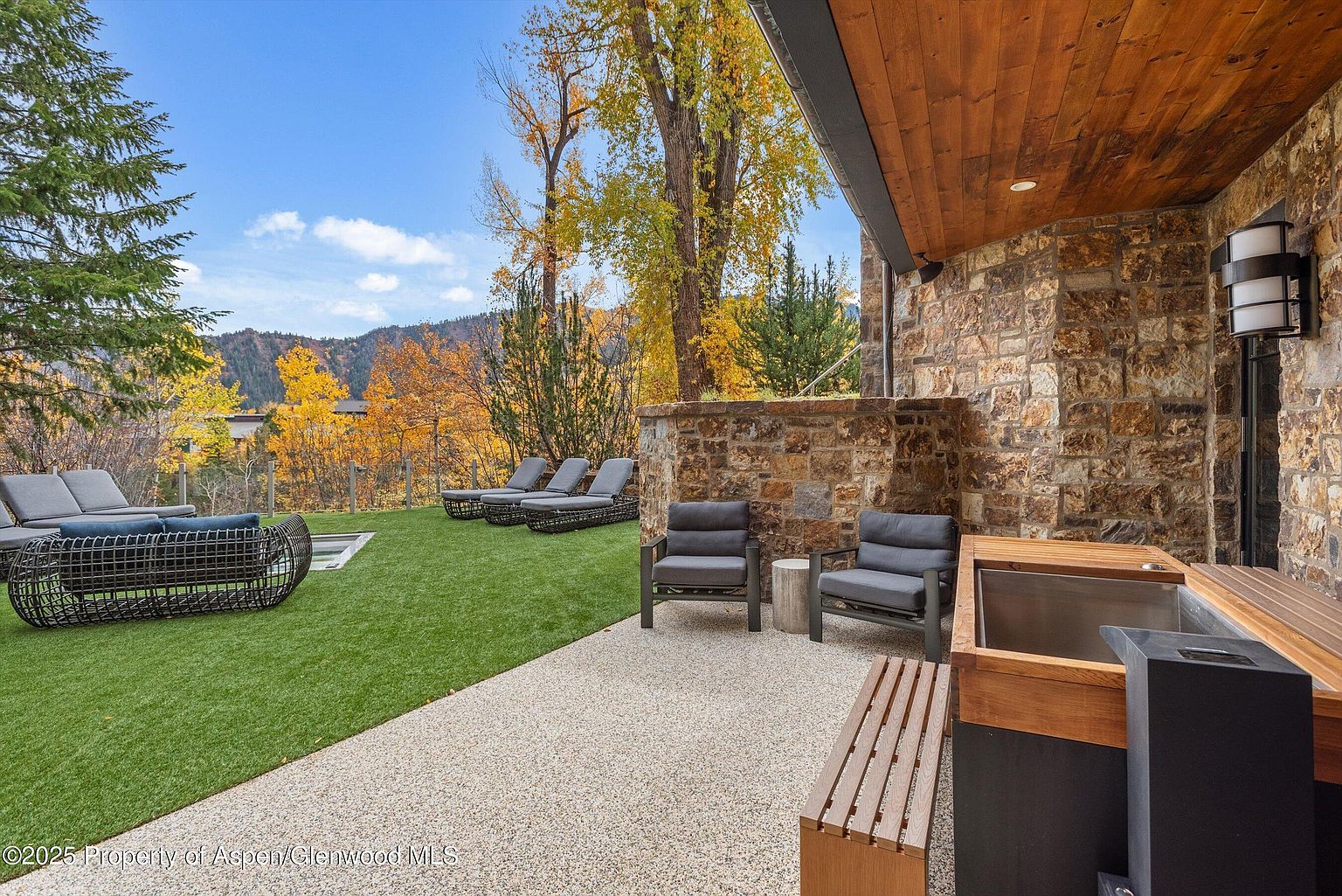 This image showcases a luxurious outdoor patio area, featuring modern lounge furniture arranged on a well-maintained lawn and a stone-paved section. The patio includes comfortable seating, a built-in hot tub, and a stone wall that adds privacy and architectural interest. The backdrop of autumn foliage and distant mountains enhances the serene and upscale ambiance, making it an ideal space for relaxation and entertainment.