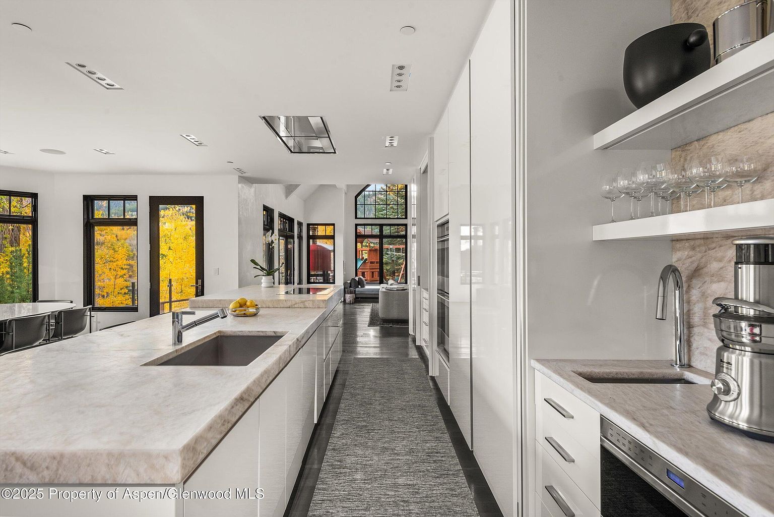 This is a bright, modern kitchen featuring a long island with a marble countertop and a stainless steel sink. The kitchen has white cabinets and appliances, with a gray runner rug adding a touch of texture. Large windows offer views of the outdoors, and open shelving displays glassware and decorative items.