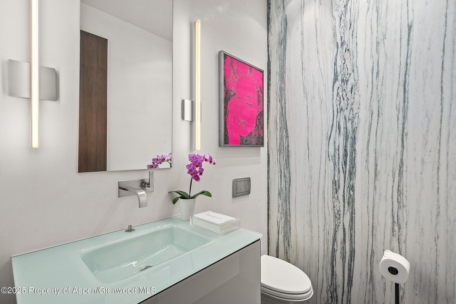 This is a modern guest bathroom featuring a unique light green glass countertop with an integrated sink. The walls are a combination of white paint and marble paneling, complemented by contemporary lighting fixtures and a framed piece of art. A toilet is visible to the right, and the overall impression is clean, stylish, and luxurious.