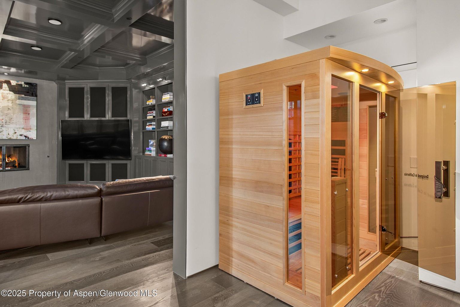 This interior shot showcases a luxurious living room with a modern sauna. The living room features a gray coffered ceiling, custom built-in shelving, a fireplace, and a comfortable leather sofa. The sauna, constructed of light wood with glass panels, adds a unique and upscale amenity to the space.