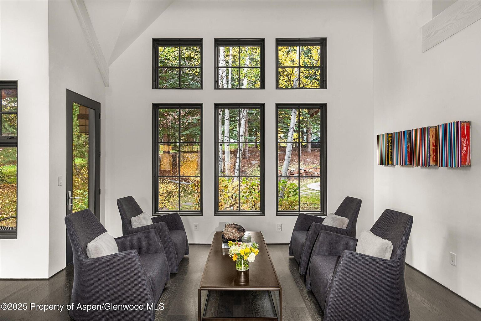 This is an interior shot of a living room featuring six armchairs arranged around a dark wood coffee table. The room is well-lit by natural light streaming through six large windows, offering a view of trees outside. The walls are white, and there is modern art on the right wall, creating a sophisticated and inviting atmosphere.