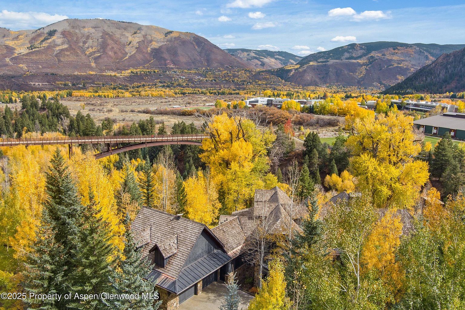 This aerial view showcases a luxurious home nestled amidst vibrant fall foliage. The house features a multi-level roofline with wood shingles and a dark exterior, complemented by surrounding evergreen trees. A pedestrian bridge spans across the landscape, leading towards buildings in the distance, with mountains providing a scenic backdrop.