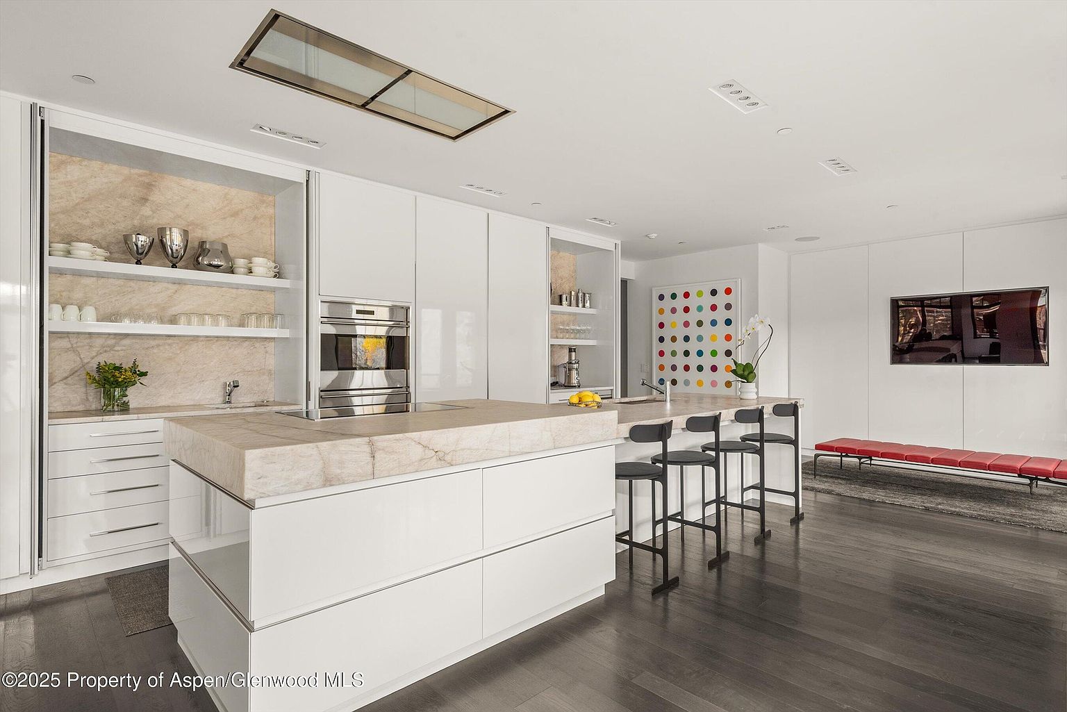 This is a modern kitchen with sleek white cabinetry and a large island featuring a marble countertop. Stainless steel appliances are integrated into the cabinetry, and the open layout includes bar seating. A colorful art piece and a television add visual interest to the space, which is illuminated by recessed lighting and a skylight.