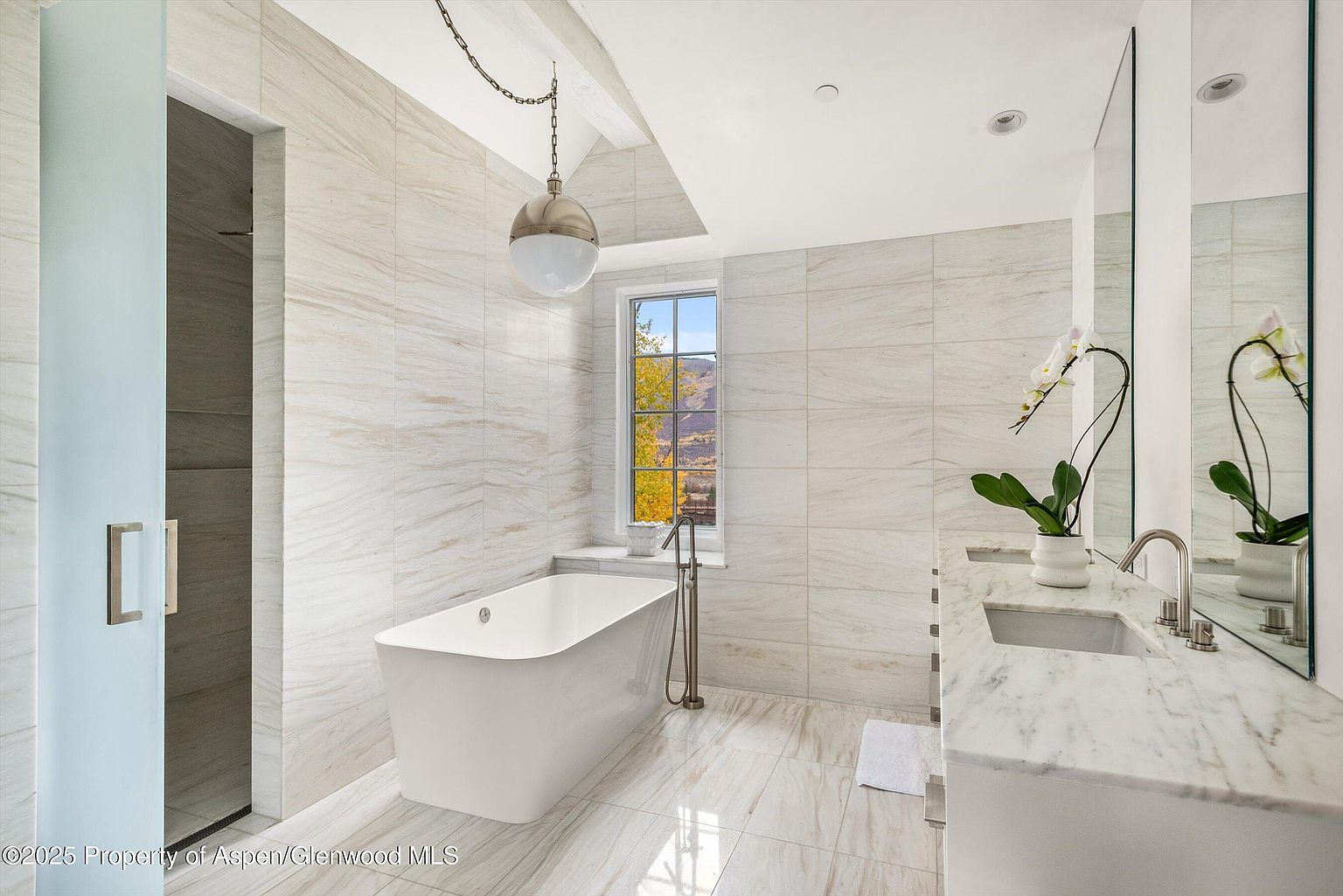 This is a bright and modern primary bathroom featuring a freestanding white bathtub positioned under a window with a view of trees. The walls and floors are covered in light-colored, horizontally-laid tiles, and a marble-topped vanity with a sink and a large mirror is visible on the right. A glass-enclosed shower is on the left, and a decorative pendant light hangs above the tub, creating a luxurious and spa-like atmosphere.