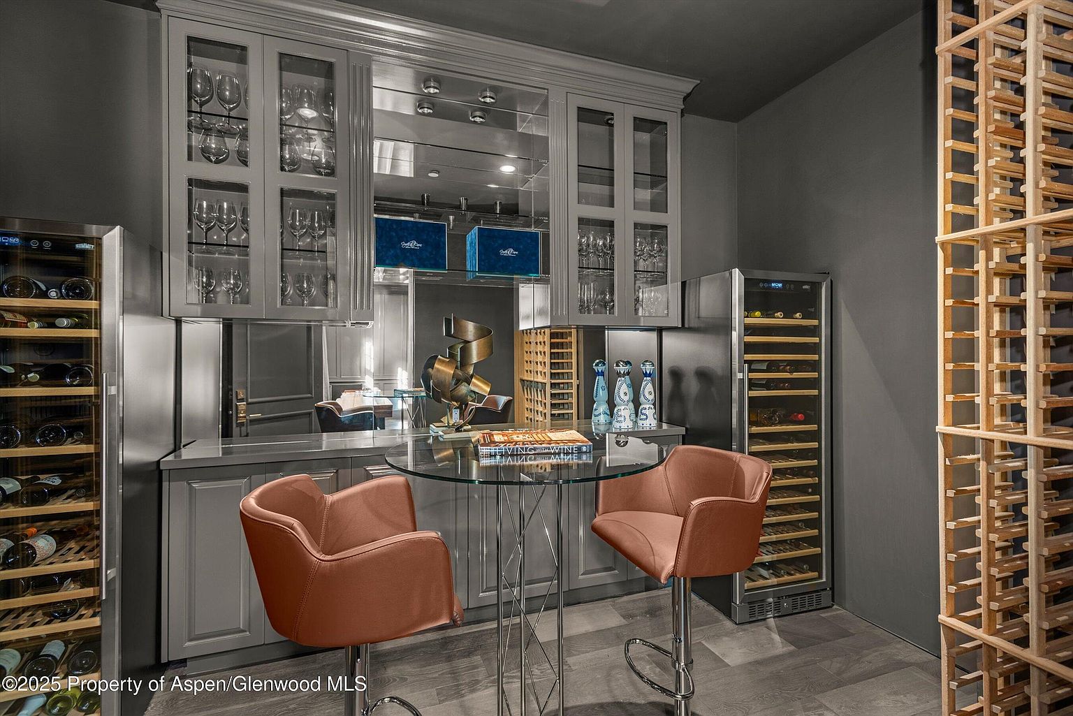 This image showcases a sophisticated wine cellar featuring custom cabinetry with glass doors displaying wine glasses and bottles. A small round glass table with two stylish chairs creates an intimate tasting area. The room is equipped with wine refrigerators and a wooden wine rack, enhancing the cellar's functionality and aesthetic appeal.