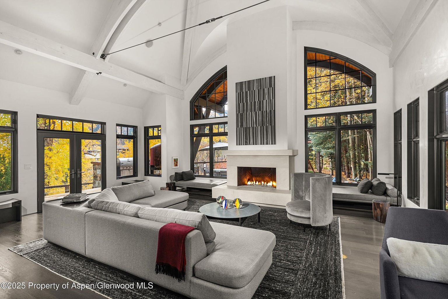 This is a spacious living room featuring high ceilings with exposed beams and large arched windows that offer views of the surrounding landscape. A large sectional sofa anchors the room, complemented by a modern fireplace and a neutral color palette. The overall impression is one of luxury and comfort, ideal for relaxation and entertaining.