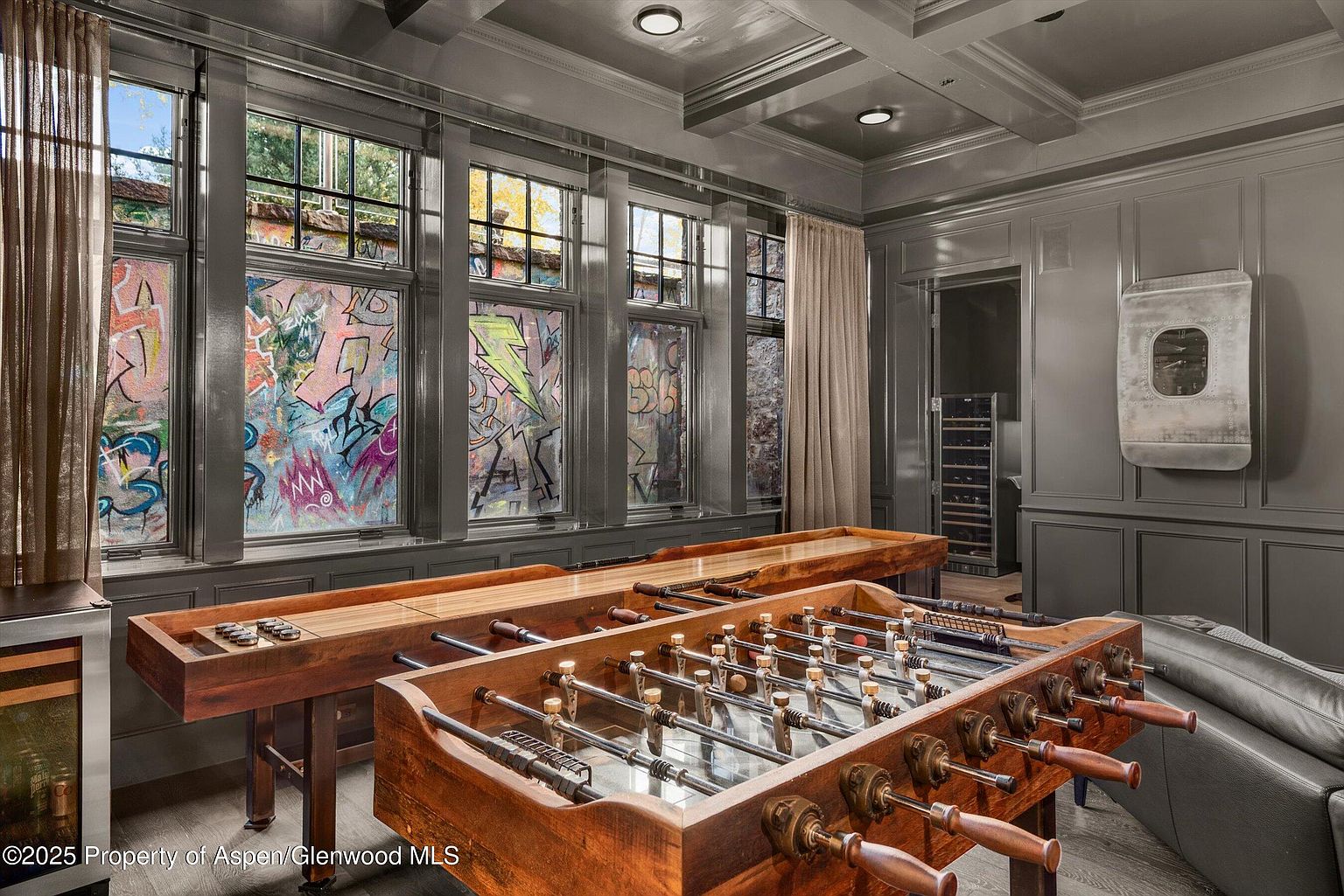 This interior shot showcases a game room featuring a foosball table and shuffleboard, creating a recreational space. The room is decorated in a modern style with gray walls and unique graffiti-style window coverings. A doorway leads to another room, possibly a wine cellar, adding to the entertainment options.