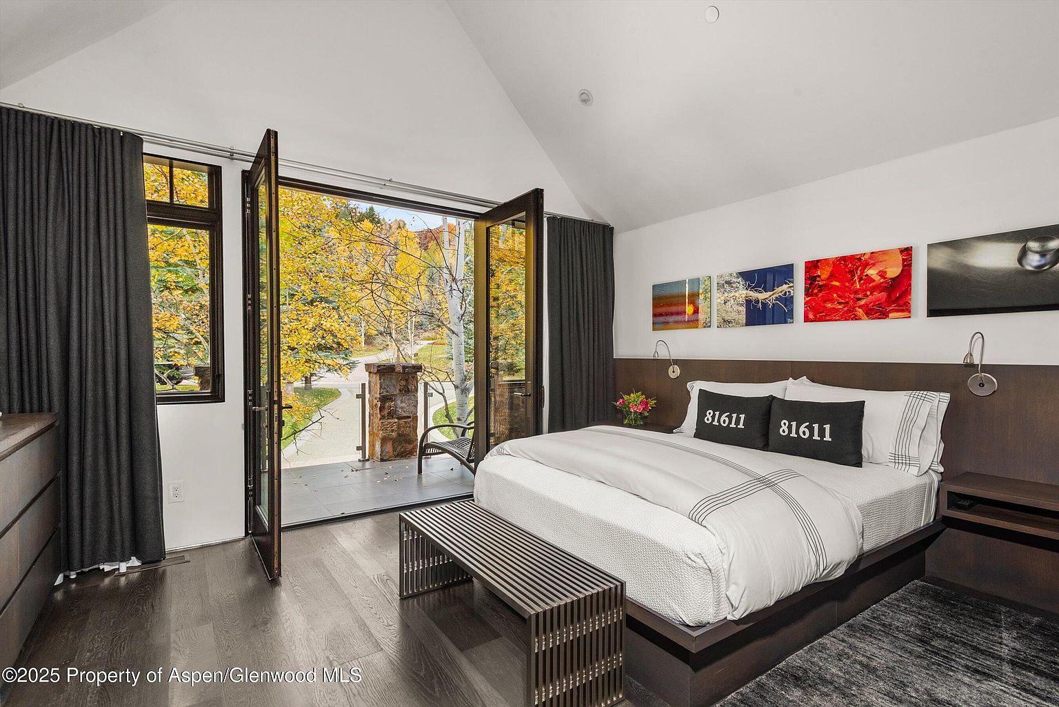 This is a primary bedroom featuring a king-size bed with a dark wood frame and a white duvet with gray stripes. The room has a modern aesthetic with artwork above the headboard and dark wood floors. Large glass doors open to a balcony with views of trees in autumn colors, creating a serene and inviting atmosphere.