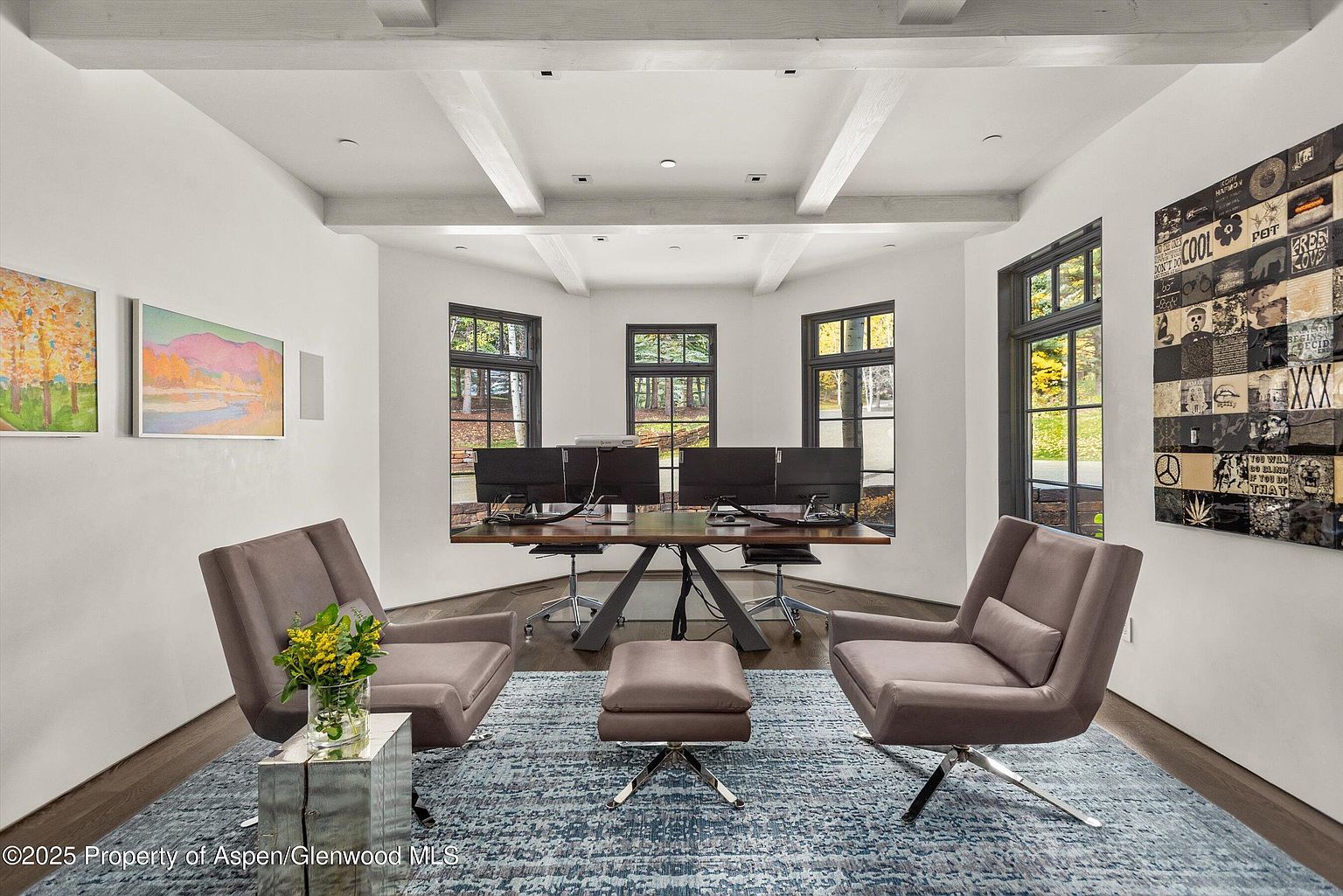 This is a well-appointed home office featuring a modern design with a focus on natural light and comfort. The room includes a large desk with multiple monitors, two stylish armchairs, and a matching ottoman, all set on a patterned blue rug. The space is bright and airy, enhanced by the white walls, exposed beams, and artwork, creating an inviting and productive work environment.