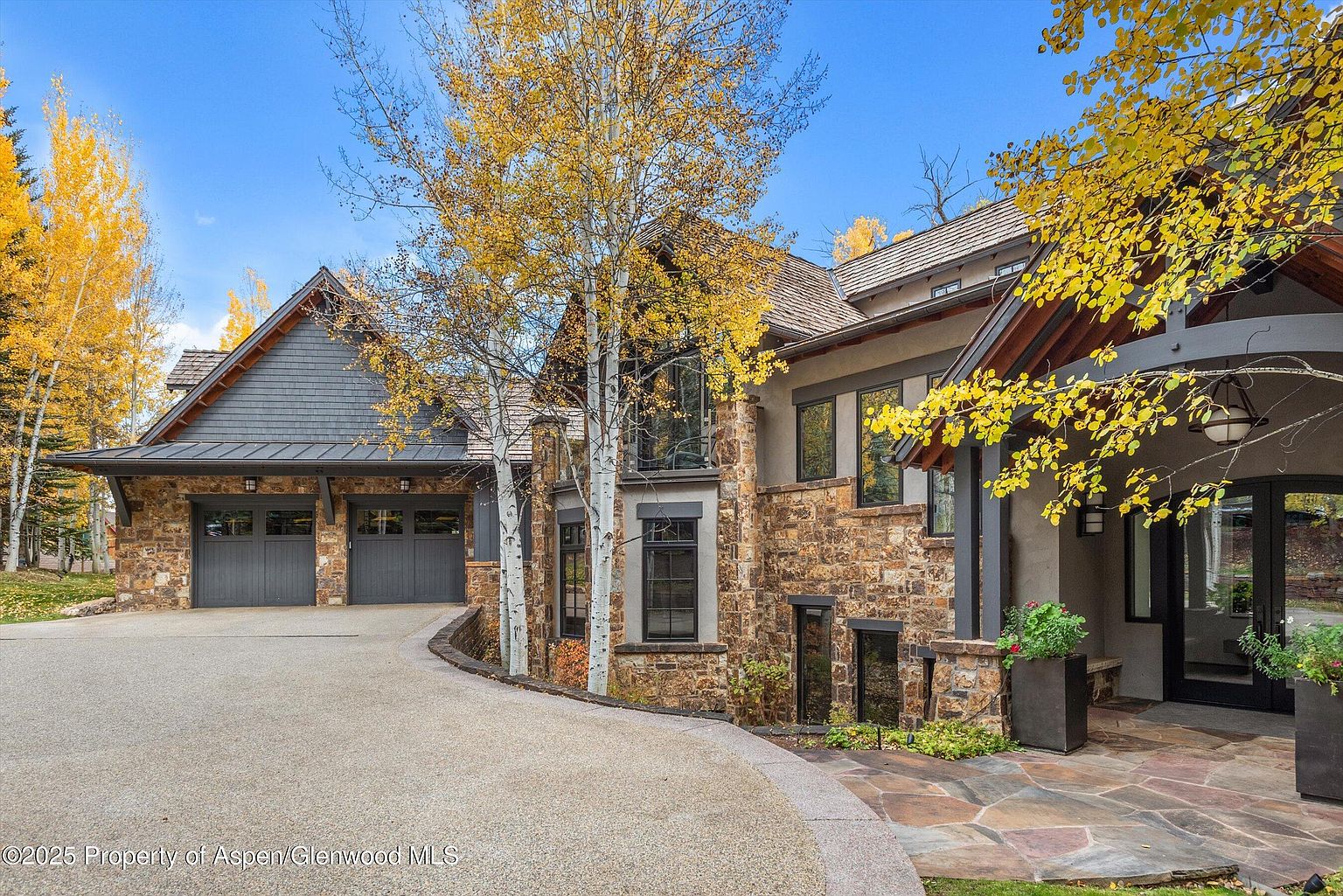This is a front exterior view of a luxurious home, showcasing a blend of stone and shingle siding. The property features a two-car garage, multiple windows, and a well-manicured driveway leading to the entrance. Mature trees with golden leaves add to the curb appeal, suggesting a serene and upscale living environment.