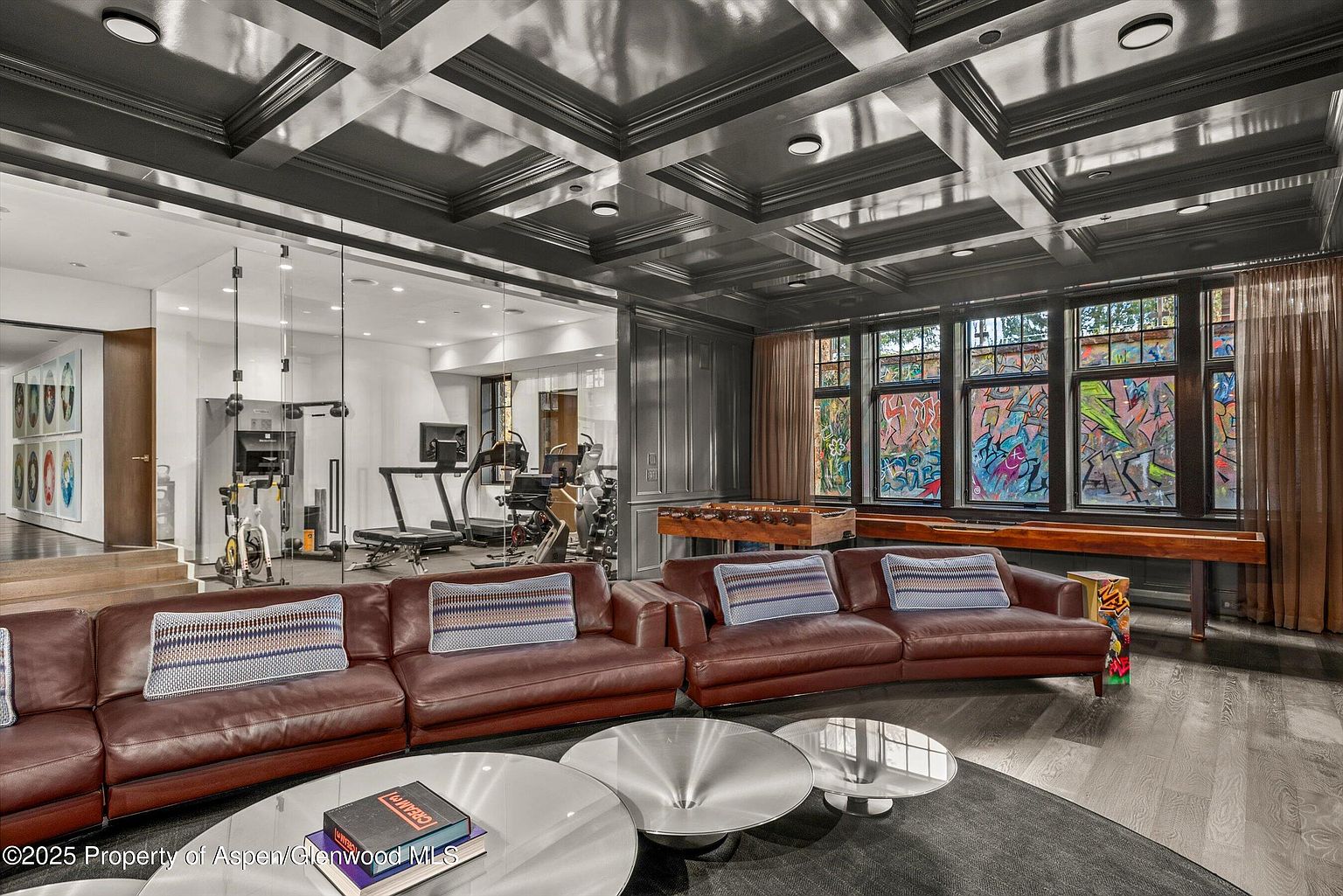 This is an interior shot of a luxurious living room featuring a curved brown leather sofa with patterned pillows, multiple round silver coffee tables on a dark rug, and a coffered ceiling with recessed lighting. The room also includes a foosball table, a shuffleboard table, and a view into a home gym through a glass wall, adding a unique entertainment and fitness aspect to the space.