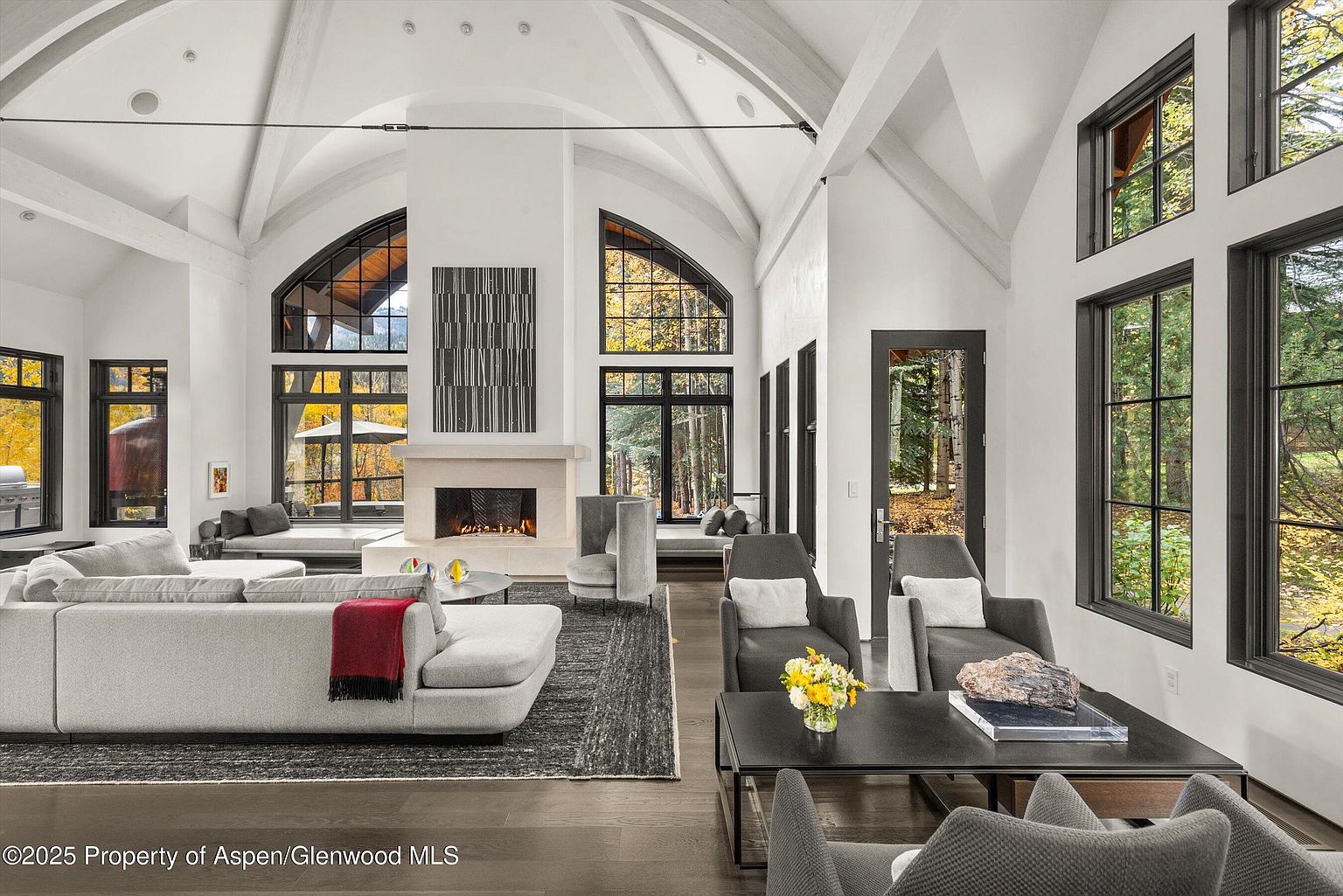 This is a spacious living room featuring high vaulted ceilings with exposed beams and large arched windows that provide ample natural light and views of the surrounding landscape. The room is furnished with a large sectional sofa, modern armchairs, and a sleek coffee table, all arranged around a contemporary fireplace. The overall design is elegant and inviting, creating a comfortable and stylish living space.