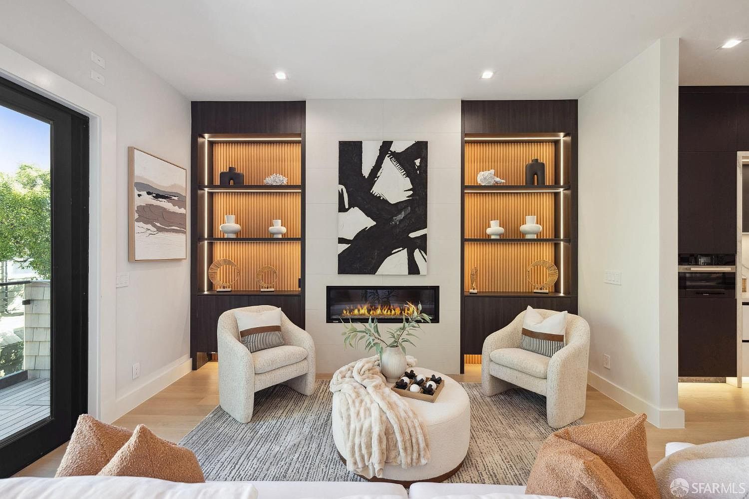 This is an interior shot of a living room featuring a modern fireplace with built-in shelving on either side. Two armchairs face the fireplace, flanking a round ottoman serving as a coffee table. The room is decorated with neutral tones and abstract art, creating a sophisticated and inviting atmosphere.