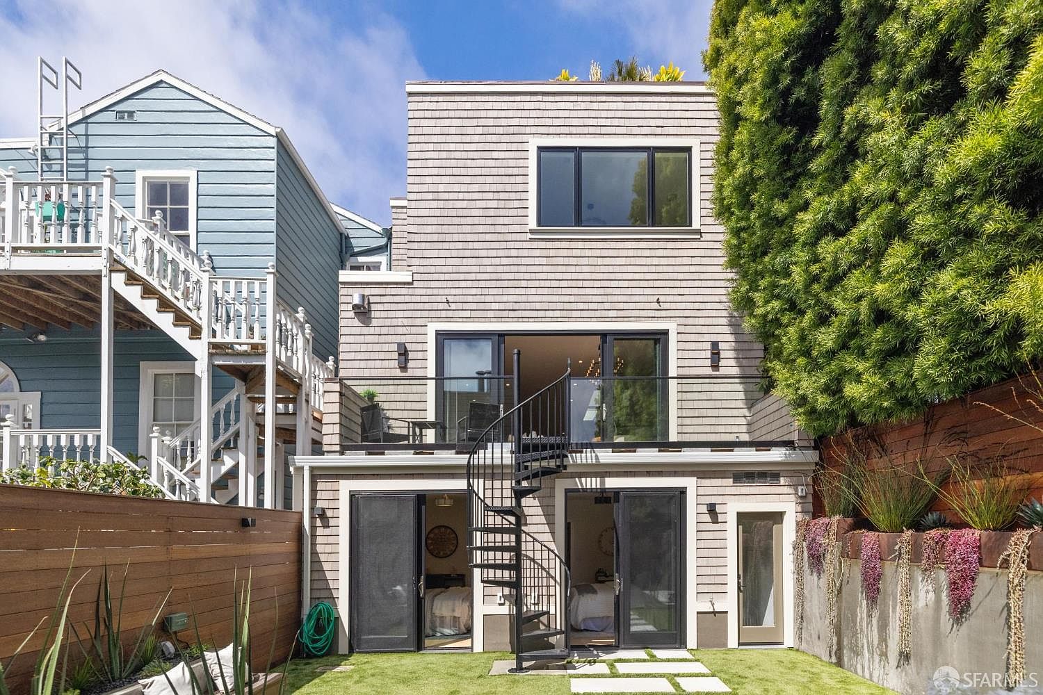 This is a rear view of a multi-story house with a cedar shingle exterior. A black spiral staircase connects the ground level to a second-story balcony. The backyard features a small lawn area and a garden bed with purple flowers, creating a private and inviting outdoor space.