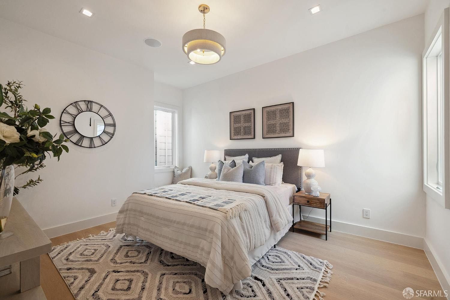 This is a well-lit bedroom featuring a queen-sized bed with a gray headboard, adorned with neutral-toned bedding and decorative pillows. Two framed art pieces hang above the bed, complemented by matching bedside lamps on a wooden nightstand. A patterned rug adds texture to the light hardwood floors, and a large clock on the wall serves as a focal point.