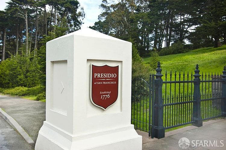 The image showcases the entrance to the Presidio of San Francisco, featuring a prominent white pillar adorned with the Presidio's emblem. A wrought-iron fence extends from the pillar, leading into a lush green landscape with mature trees in the background. The scene conveys a sense of history and prestige, highlighting the property's unique location and character.