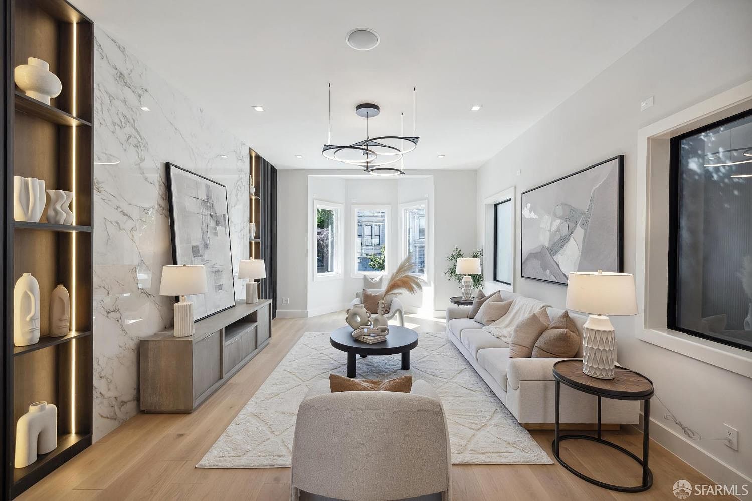 This is a bright and airy living room featuring a modern design with a neutral color palette. The room is well-lit with natural light from the bay windows and complemented by stylish lighting fixtures. Key features include a marble accent wall, a sleek entertainment center, a comfortable sofa, and a round coffee table, creating an inviting and sophisticated space.