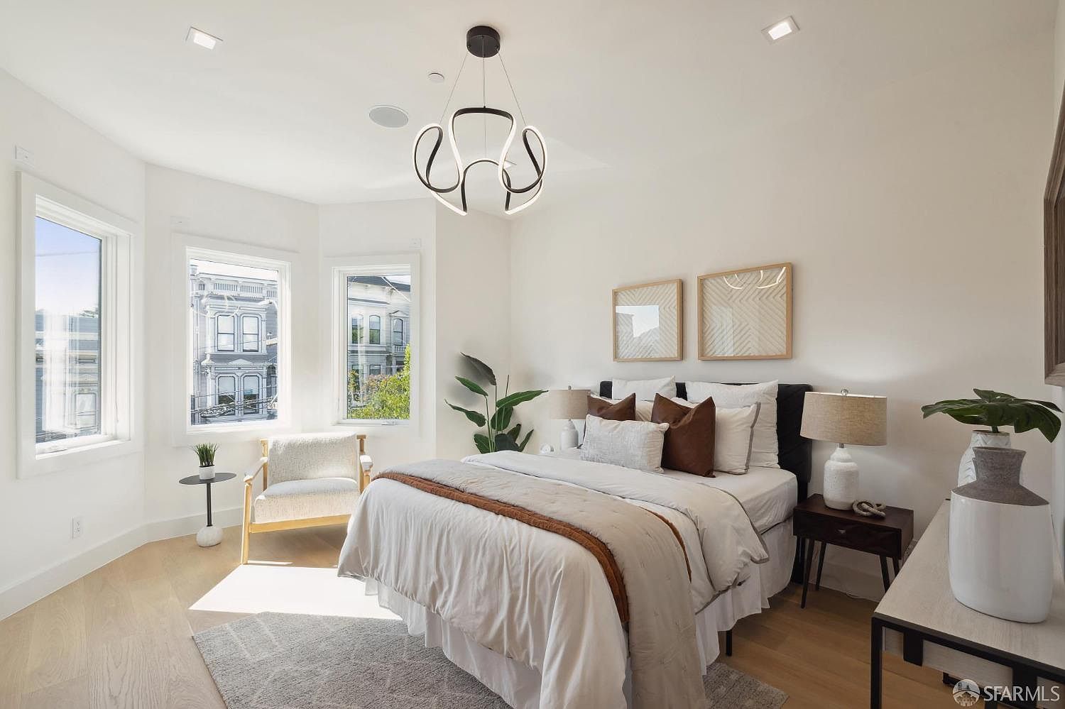 This is a bright and airy primary bedroom featuring a large bed with neutral bedding and decorative pillows. The room has hardwood floors, white walls, and a modern chandelier. Natural light floods the space through a bay window, creating a serene and inviting atmosphere.