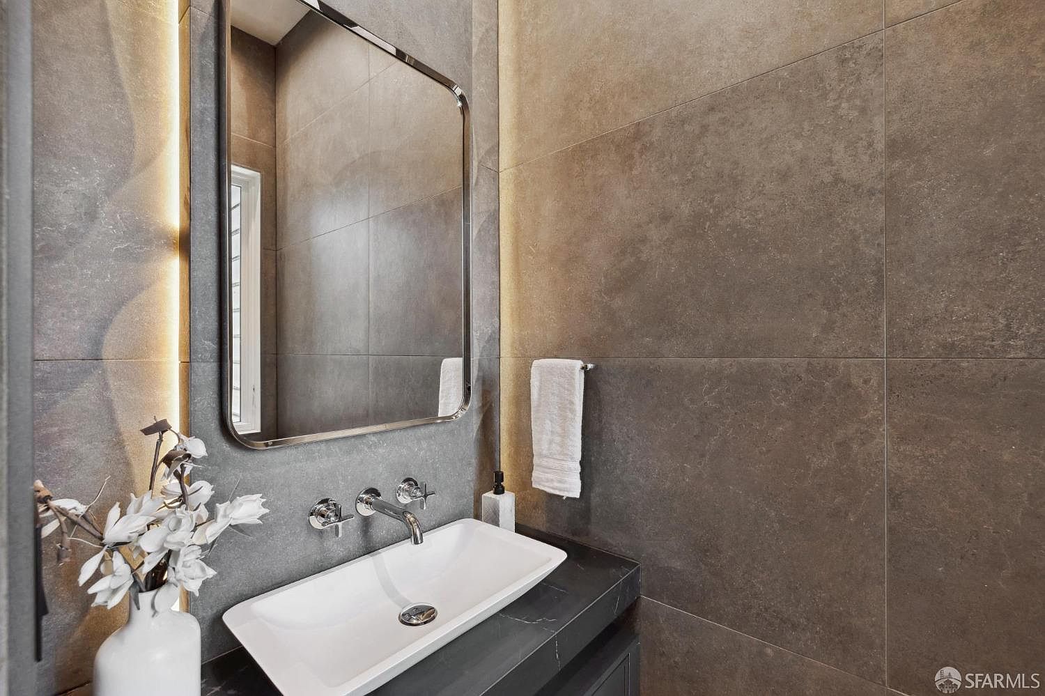 This is a modern bathroom featuring a rectangular white sink atop a dark countertop, complemented by sleek chrome fixtures. A large, vertically oriented mirror with rounded corners reflects the space, while a neutral-toned tiled wall provides a sophisticated backdrop. A white towel hangs neatly on a chrome bar, adding a touch of elegance.