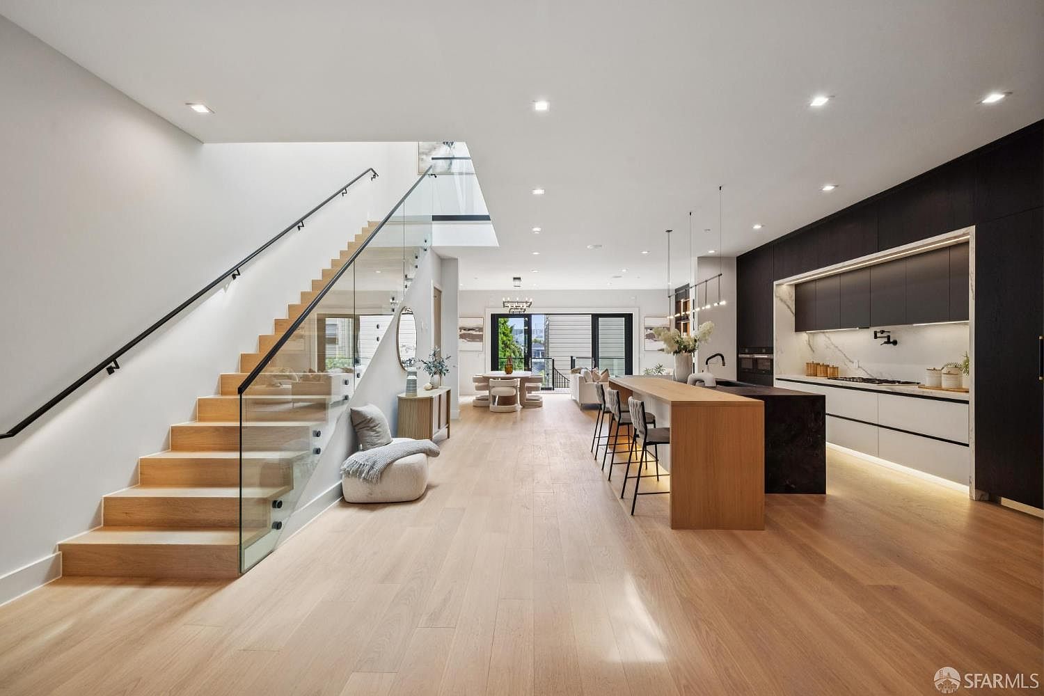 This is a bright, open-concept kitchen and living area featuring modern design elements. The kitchen boasts sleek, two-tone cabinetry, a large island with bar seating, and high-end appliances. The living area includes a staircase with glass railings and a comfortable seating area, all contributing to a luxurious and inviting atmosphere.