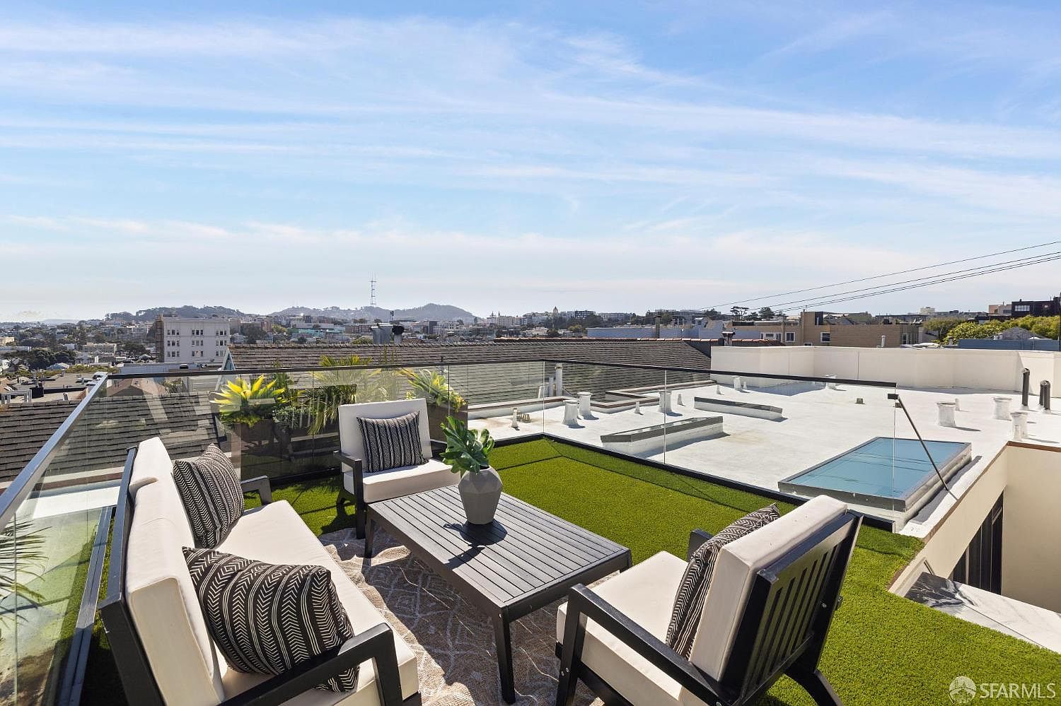 This is a high-angle shot of a luxurious rooftop patio. The patio features modern outdoor furniture, including a sofa and chairs with neutral cushions and patterned pillows, arranged around a dark wood coffee table. Artificial turf adds a touch of greenery, and a small pool is visible in the background, all set against a panoramic city view under a clear blue sky.
