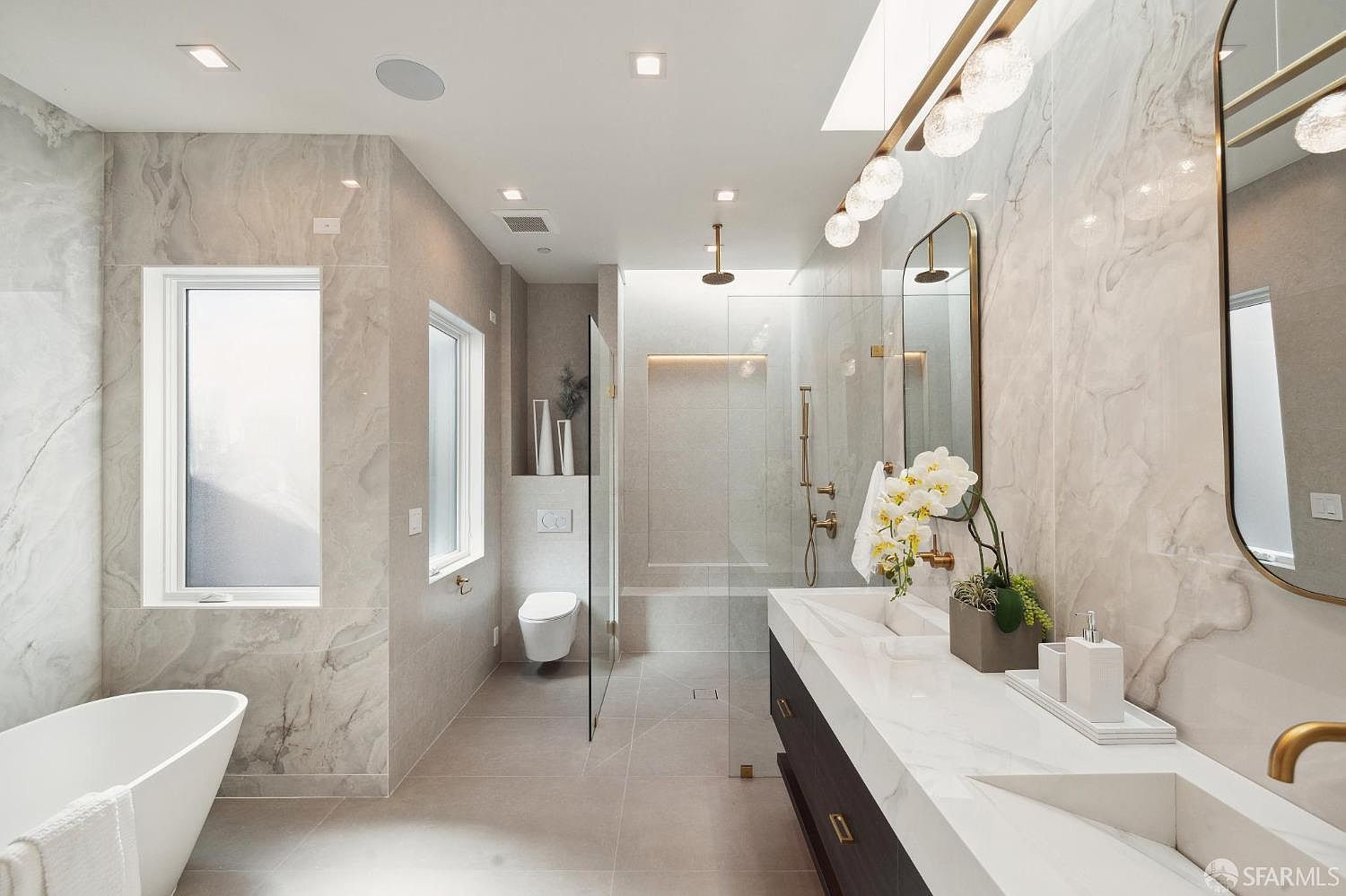This is a luxurious primary bathroom featuring marble-like tile walls and flooring. The room includes a freestanding bathtub, a floating double vanity with a white countertop and dark wood cabinets, and a glass-enclosed shower with gold fixtures. Natural light floods the space through a skylight and windows, creating a bright and airy atmosphere.