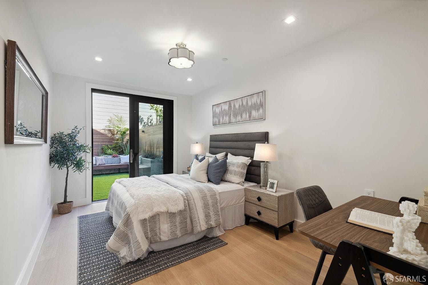 This is a bright and stylish bedroom featuring a bed with a gray headboard, striped bedding, and a fluffy throw. A sliding glass door leads to a small outdoor patio with artificial turf and a built-in bench. The room also includes a wooden desk and chair, creating a functional workspace.