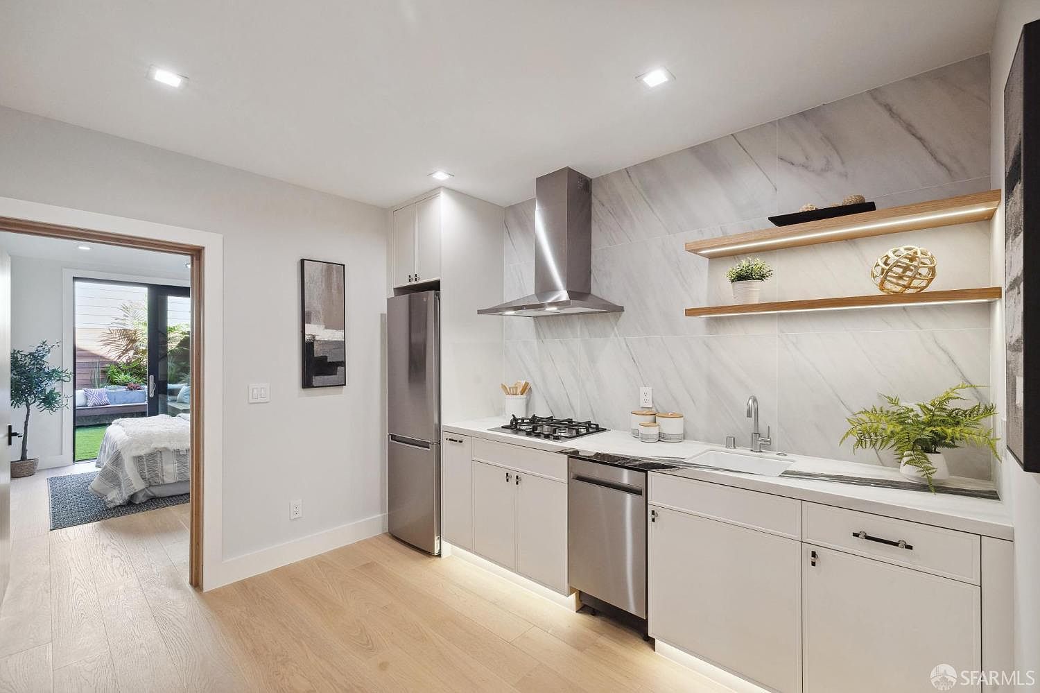 This is a well-lit, modern kitchen featuring light wood flooring and white cabinetry. Stainless steel appliances, including a refrigerator, dishwasher, and range hood, add a sleek touch. Open wooden shelves with integrated lighting provide a stylish display area, while a doorway leads to an outdoor patio area, creating a seamless indoor-outdoor flow.