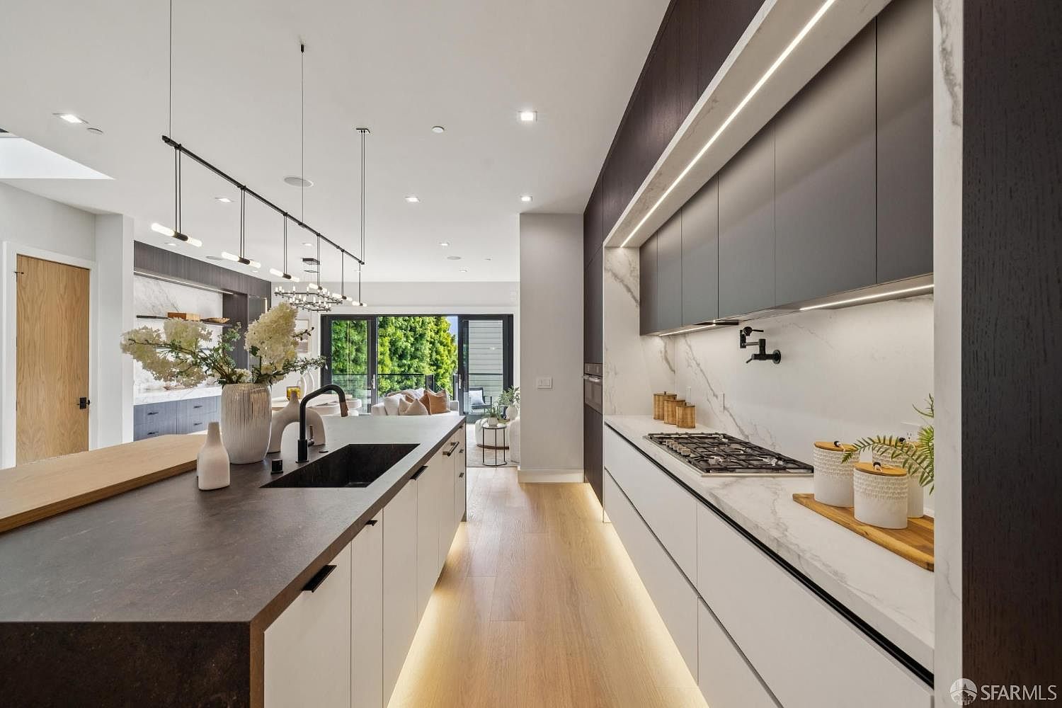 This is a modern kitchen with a long island featuring a dark countertop and white cabinetry. The kitchen also includes sleek, gray upper cabinets with integrated lighting and a marble backsplash. The open layout leads to an outdoor living space, creating a seamless transition between indoor and outdoor living.