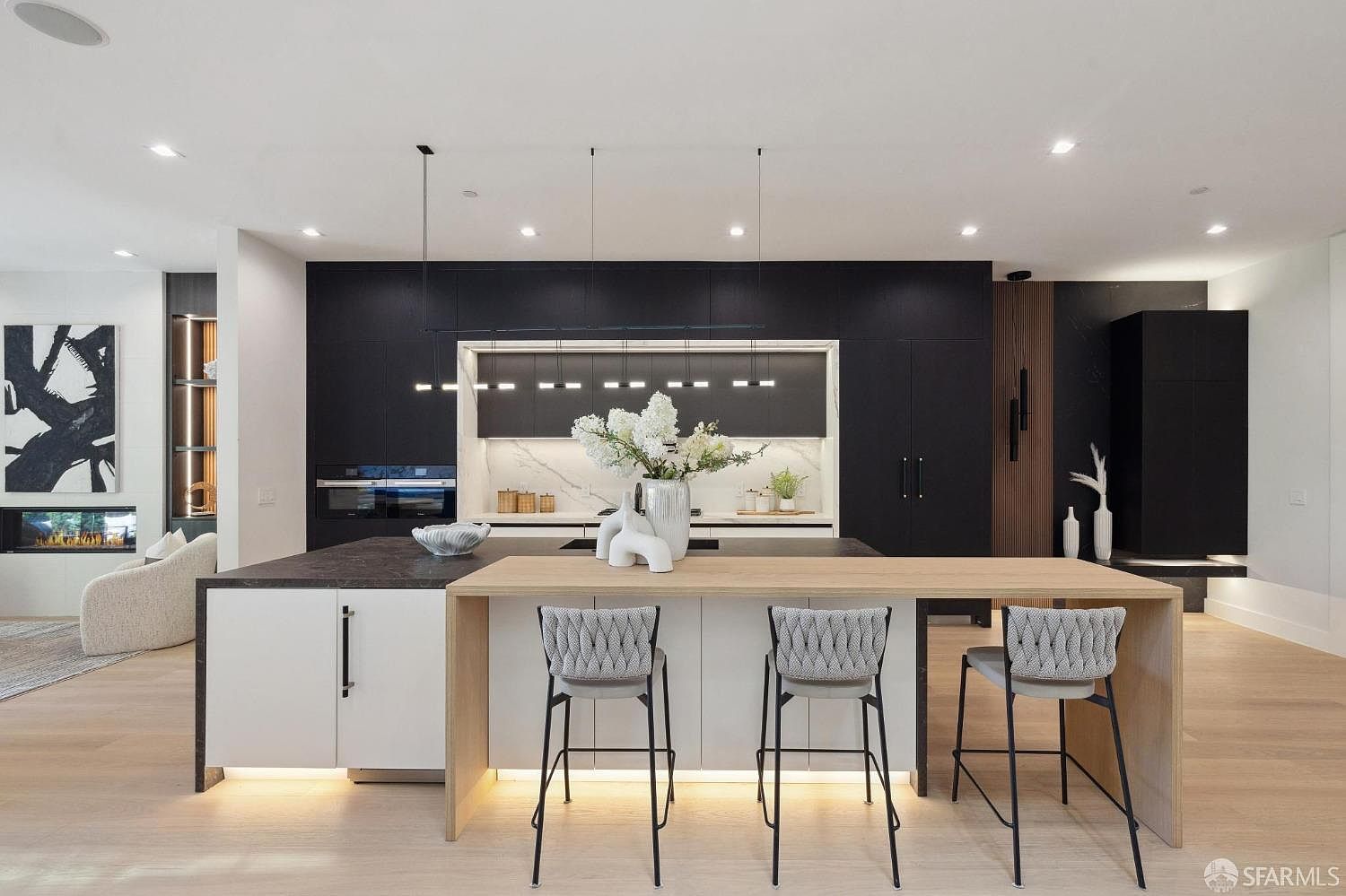 This is a modern kitchen featuring a large island with seating, sleek black cabinetry, and a contrasting white backsplash. The island has a light wood extension, and the space is illuminated by modern pendant lighting and under-cabinet lighting. The overall impression is one of sophisticated design and functionality.